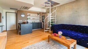 A modern living room with a wooden floor, a blue velvet sofa, a wooden coffee table, and a spiral staircase leading to a kitchen area. - Fiftyfive-one Zuretti (Milano)