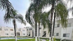 A stone barbecue grill with a metal grate, surrounded by a stone wall, set in a courtyard with palm trees and a pool. - Departamento en Fraccionamiento Seguro

 (Yautepec de Zaragoza)