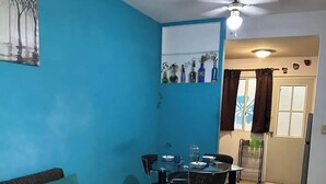 A room with a blue wall, a ceiling fan, a dining table with chairs, a sofa with teal cushions, and a door leading to another room. - Departamento en Fraccionamiento Seguro

 (Yautepec de Zaragoza)