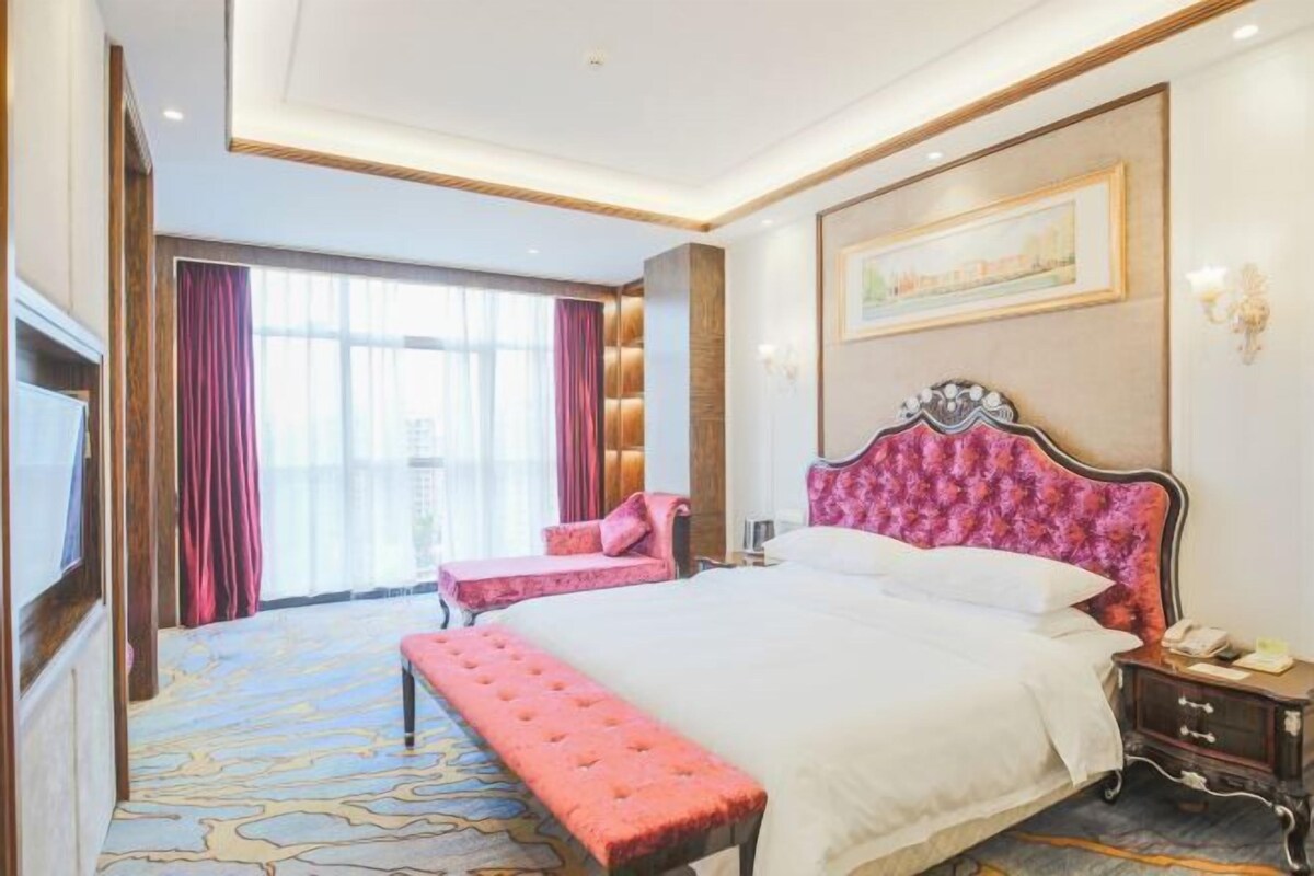 A hotel room with a large bed, a bench, a bedside table, a TV, and a window with curtains.
