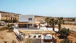 A modern villa with a stone facade, a wooden pergola, and a pool, surrounded by a dry landscape and palm trees. - Diamond of Cyclades (Kastraki)