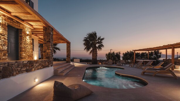A pool area with lounge chairs, a stone wall, and a wooden pergola. - Diamond of Cyclades (Kastraki)