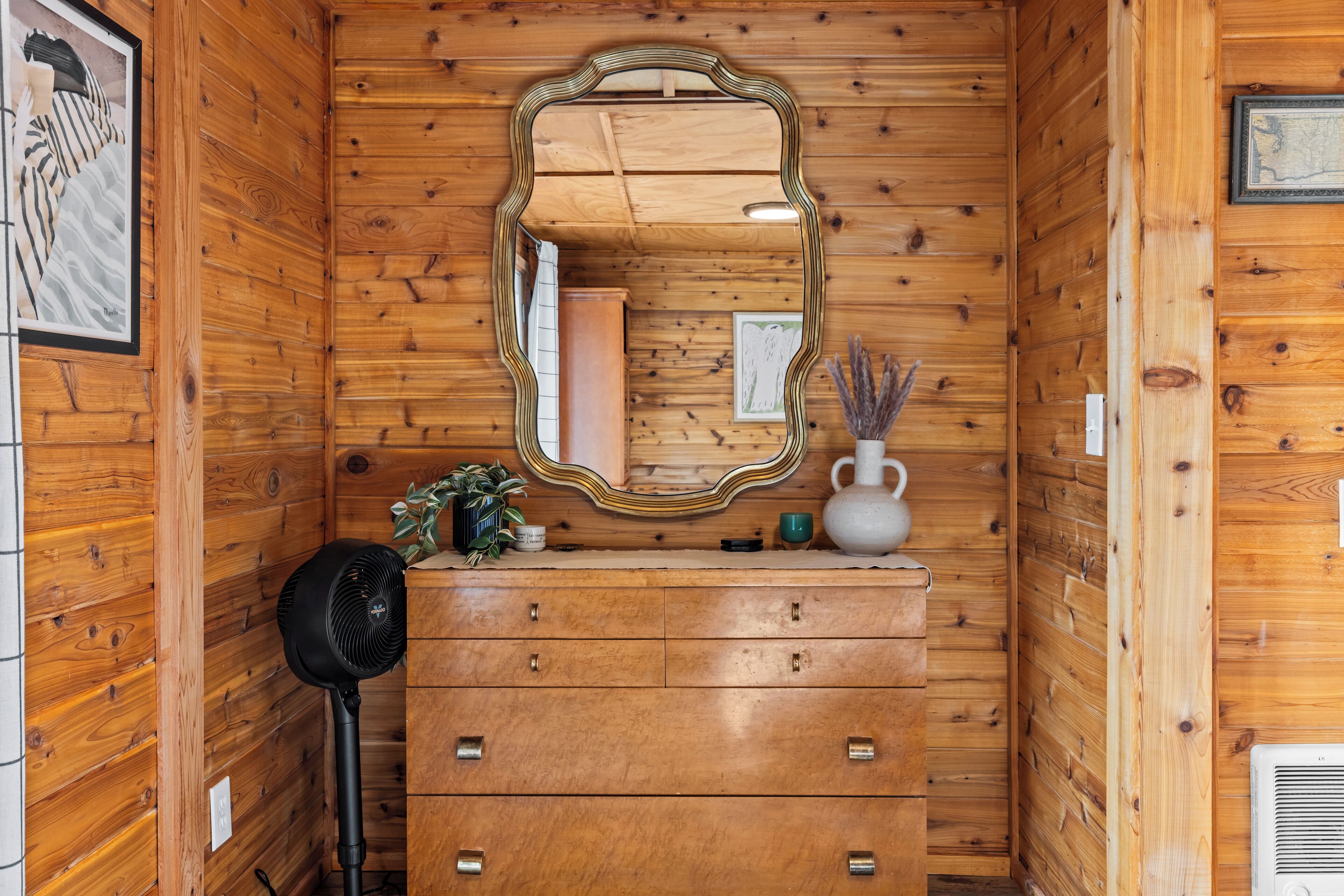 Interno di una casetta di legno con un comò, uno specchio e una ventola.