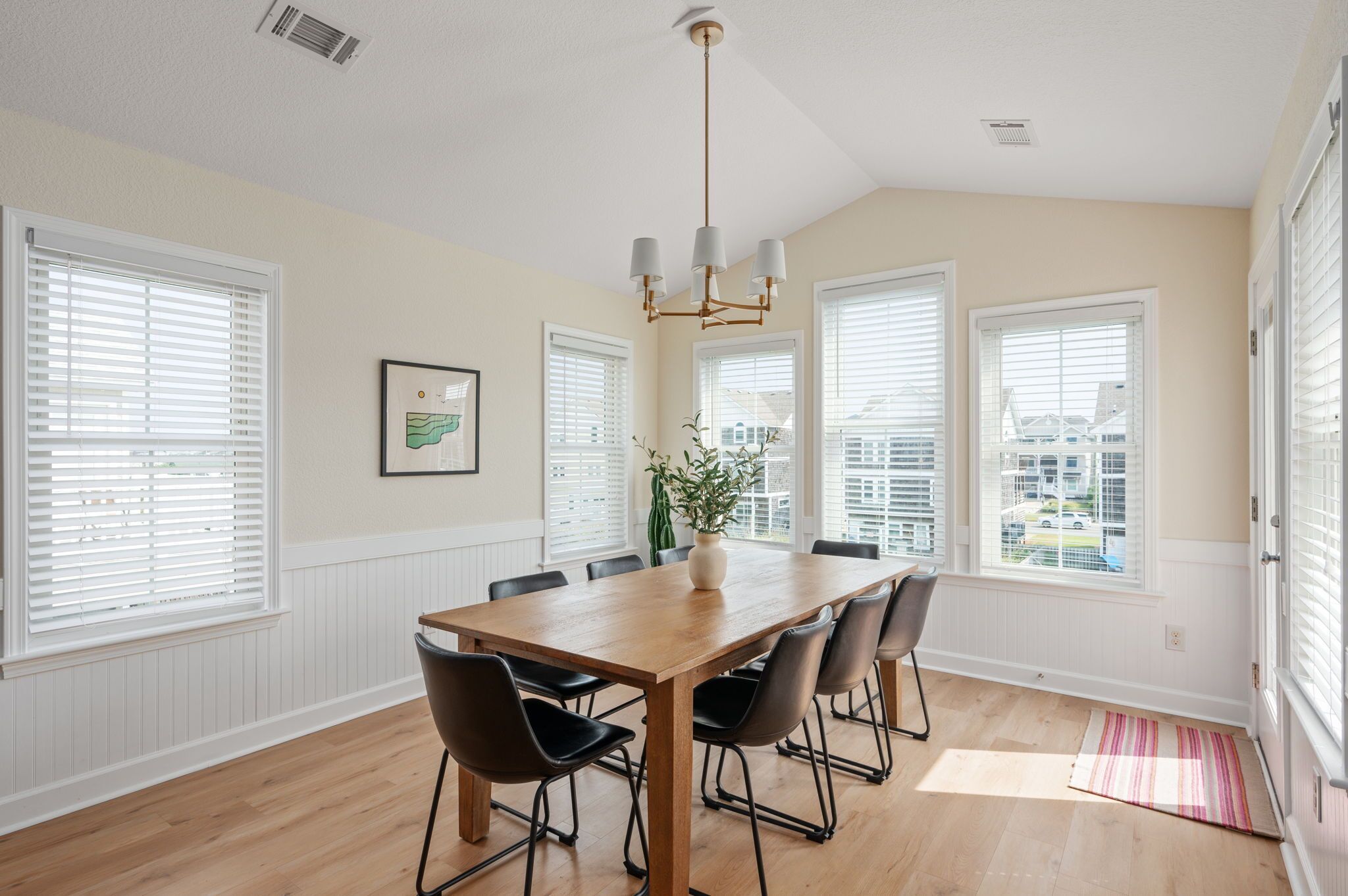 A dining area with a wooden table, six chairs, a chandelier, and large windows with blinds.