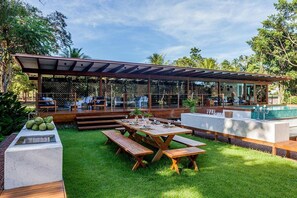 Un espace de restauration extérieur moderne, avec des tables et des bancs en bois, une piscine et un coin salon abrité.