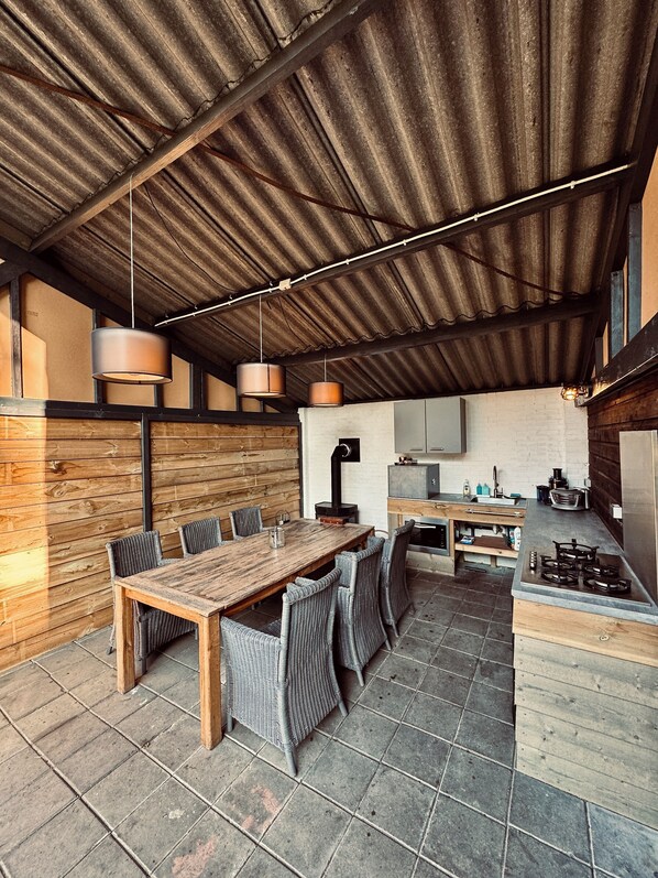 An outdoor dining area with a wooden table and chairs, a kitchen with a stove and sink, and a corrugated metal roof.