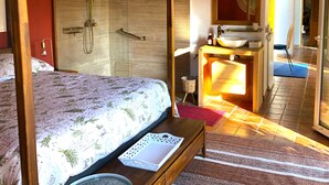 A bedroom with a bed, a wooden nightstand, a bedside table, a large window, a bathroom with a sink and mirror, and a shower.