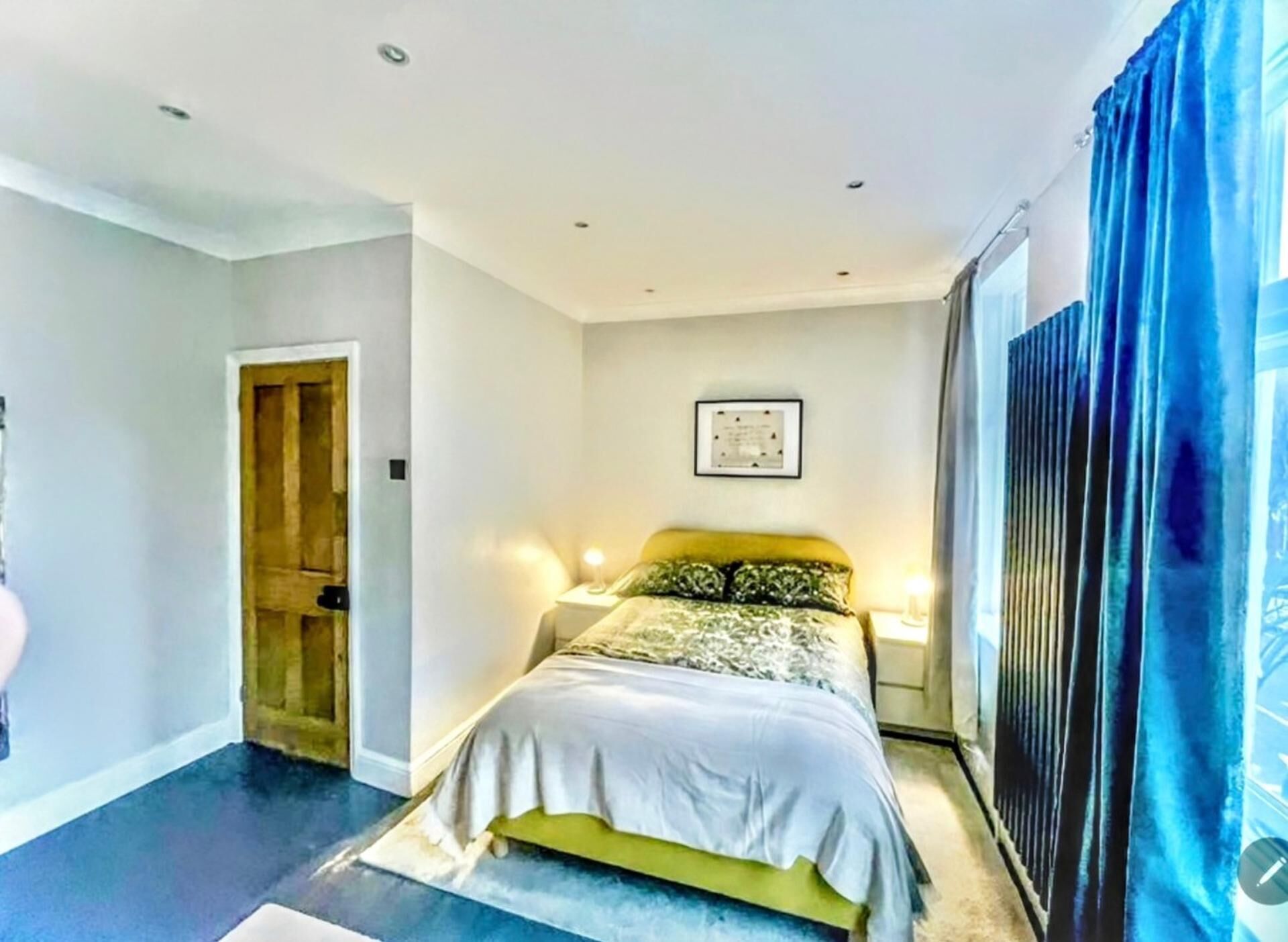 A bedroom with a bed, bedside tables, a framed picture on the wall, and a large window with curtains.