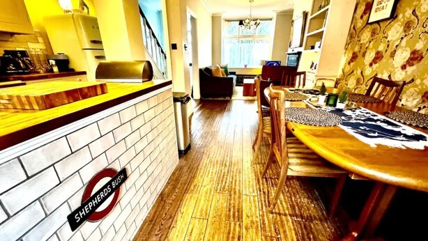 A dining area with a wooden table and chairs, a brick wall with a 'Shepherds Bus' sign, and a staircase leading to another room. - Shepherds Bush W12 Magical Townhouse nr Westfield (London)