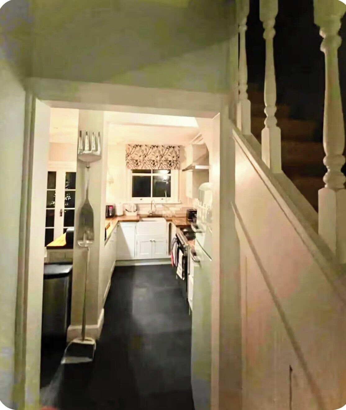 A narrow hallway leading to a kitchen with white cabinets, a window with a patterned curtain, and a staircase on the right.