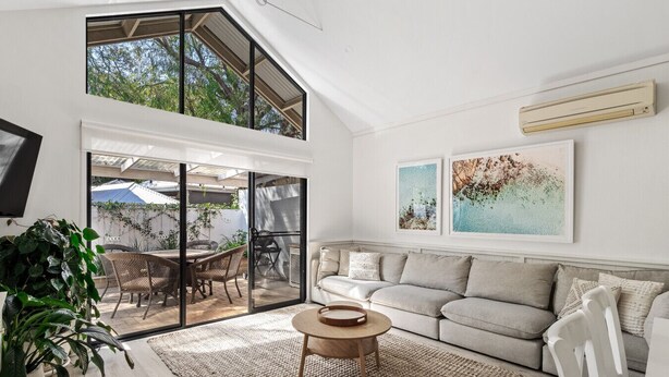 A spacious living room with a high ceiling, a sliding glass door leading to an outdoor patio, a comfortable sofa set, and two framed artworks on the wall.