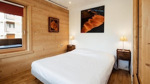 A bedroom with a wooden wall, a large bed, two bedside tables with lamps, a window, and a painting on the wall.