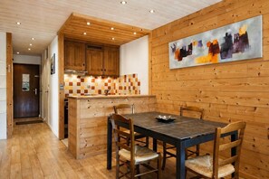 A wooden interior with a dining table, chairs, a kitchen area, and a wooden wall with a painting.