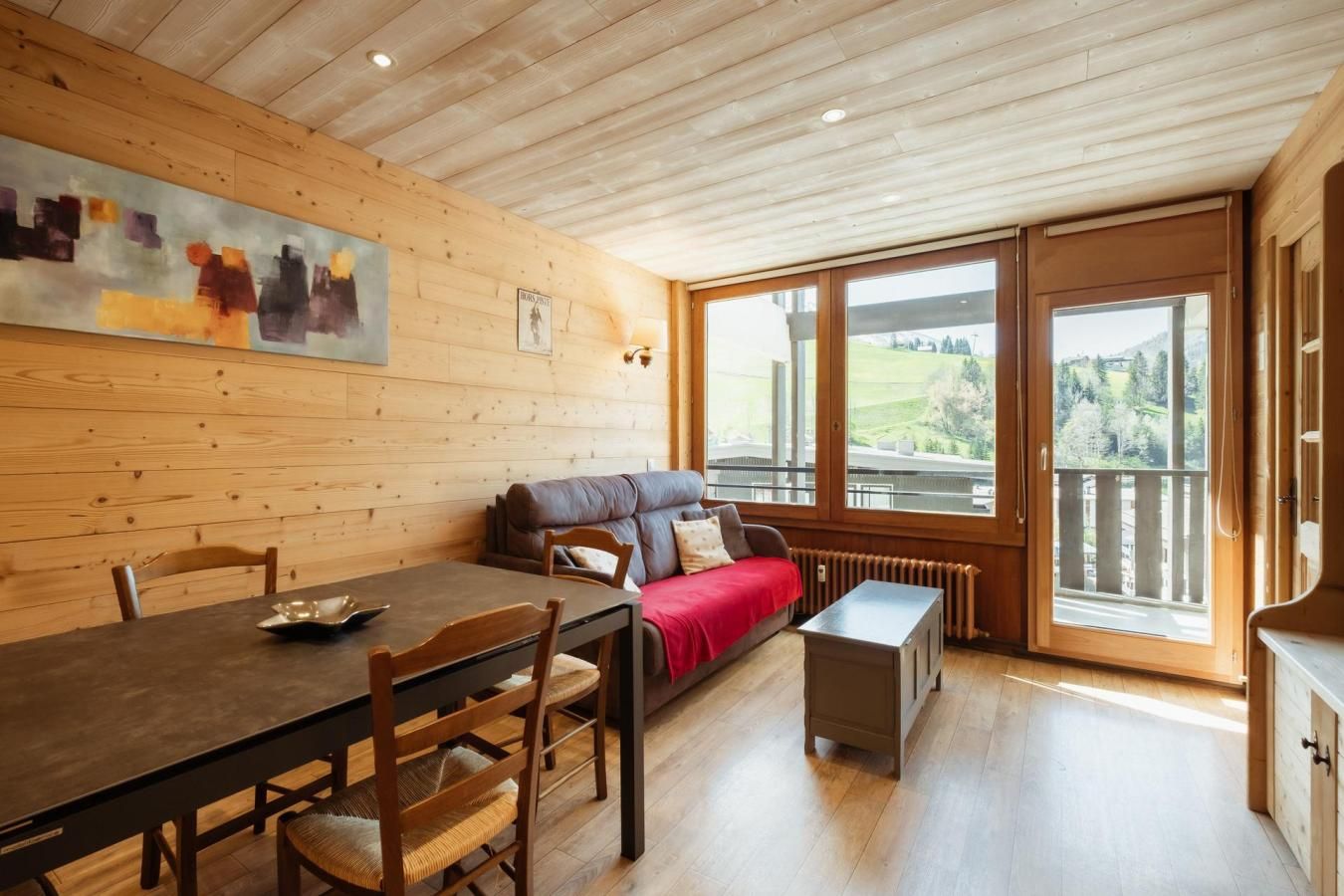 A cozy room with a wooden ceiling, a dining table with chairs, a sofa with a red blanket, and large windows offering a view of the outdoors.