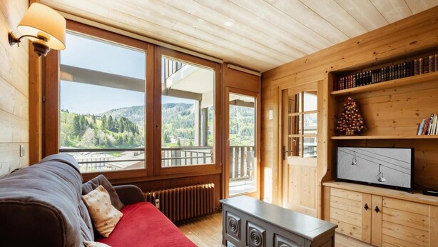 A cozy room with a wooden interior, a large window offering a scenic view, a comfortable sofa, and a television on a wooden cabinet.