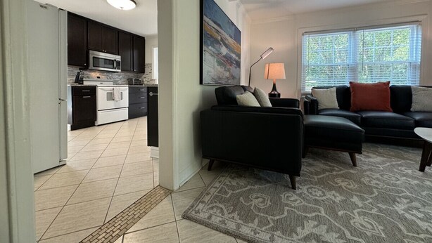A modern kitchen with dark cabinets and stainless steel appliances, a black leather sofa with cushions, a patterned rug, and a large window with blinds. - House at Guilford Hills (Greensboro)