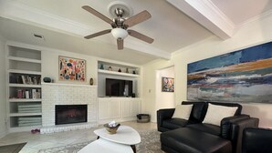 A living room with a fireplace, black leather sofa, white coffee table, and a ceiling fan. - House at Guilford Hills (Greensboro)