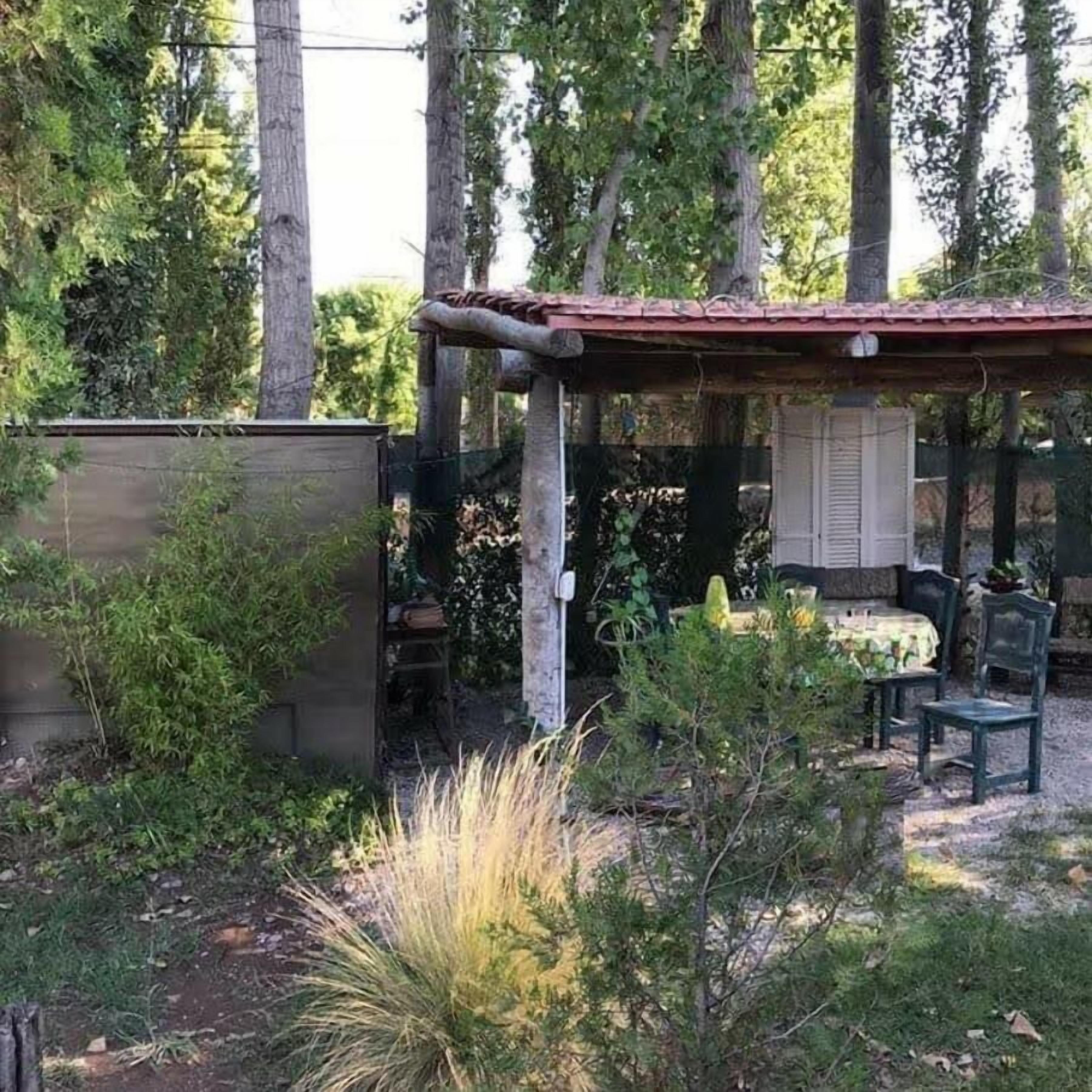 A covered outdoor seating area with a table and chairs, surrounded by trees and vegetation.