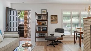 Un salon comprenant un canapé, une table basse en verre, un fauteuil de bureau noir, une bibliothèque et une grande fenêtre avec des stores.