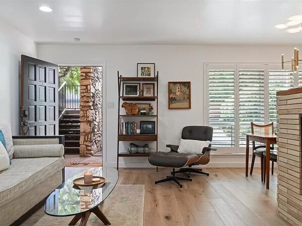 A living room with a sofa, a glass coffee table, a black office chair, a bookshelf, and a large window with blinds.