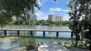 Un parc au bord de la rivière, avec une table et des bancs en pierre, un pont et des bâtiments en arrière-plan.