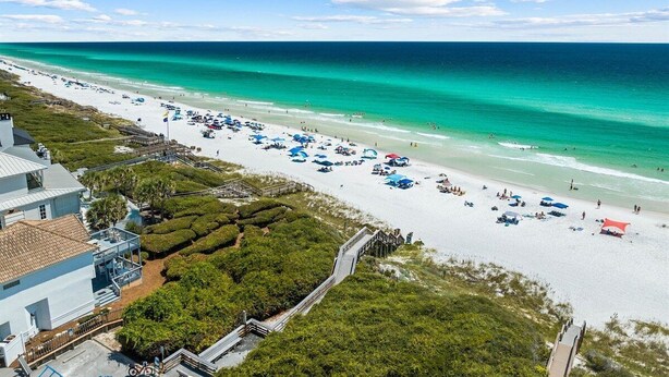 A beach with a long stretch of white sand, a clear blue ocean, and a few buildings in the background.