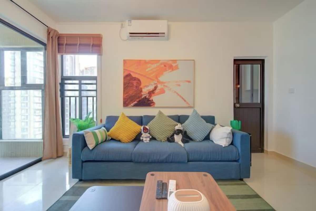 Een woonkamer met een blauwe bank, kleurrijke kussens, een houten salontafel, een afstandsbediening en een wandairconditioner.