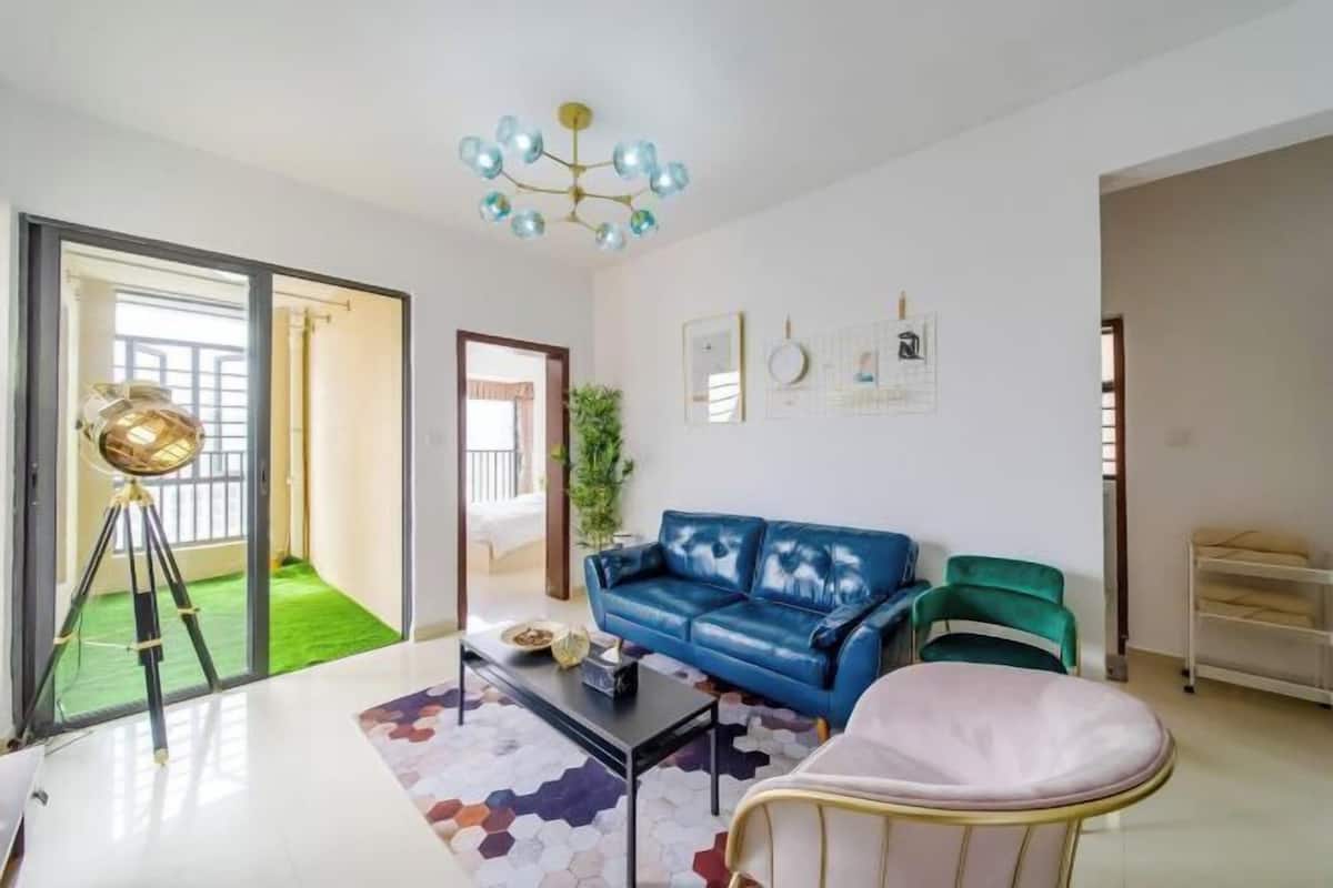 Een woonkamer met een blauwe bank, een groene fauteuil, een witte slaapstoel, een zwarte salontafel en een bijzondere plafondlamp.