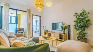 Un salon moderne avec un canapé, un pouf et un téléviseur posé sur un meuble en bois. On y trouve une grande fenêtre avec des rideaux, une plante et un luminaire suspendu.