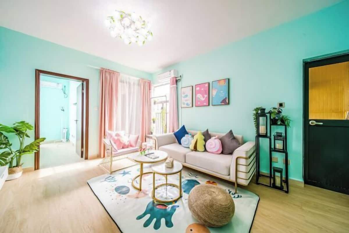 Een woonkamer met een bank, salontafel en decoratieve kussens. Er is een deur aan de rechterkant en een raam met gordijnen.