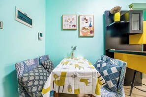 A dining area with a table covered by a patterned tablecloth, two framed illustrations on the wall, and a yellow cabinet with a plant on top. - Dream House (Zhuhai)