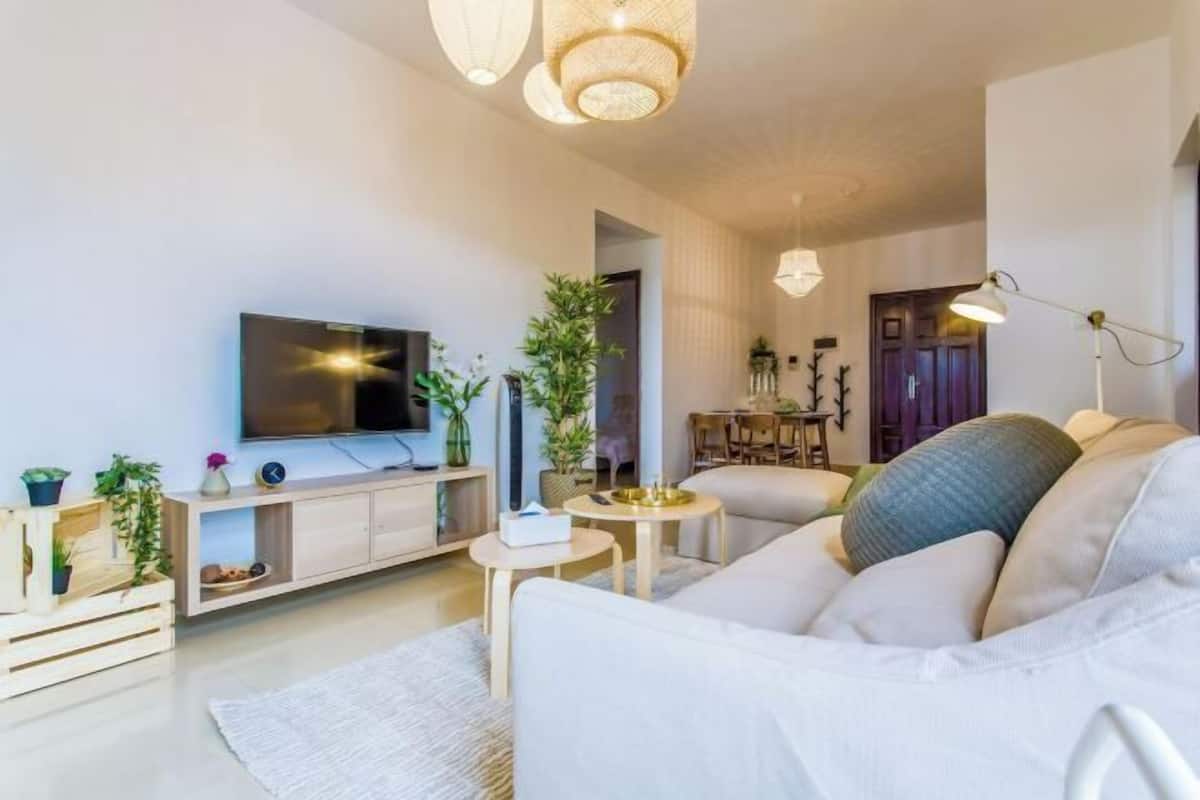 Een moderne woonkamer met een flatscreen televisie, een bank, een klein rond tafeltje en decoratieve planten.