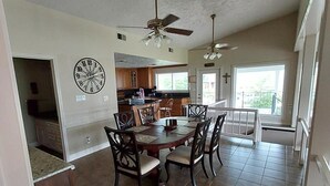 Un grand espace à manger avec une table et des chaises en bois, un ventilateur de plafond et une cuisine en arrière-plan.