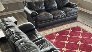 Un salon avec un canapé en cuir, un tapis à motifs rouges et de grandes fenêtres équipées de stores.