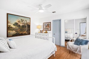 Ein Schlafzimmer mit einem großen Bett, einem Deckenventilator, einem Gemälde, einer Kommode und einer Tür zu einem anderen Raum.