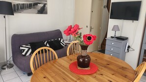 A dining area with a wooden table, chairs, a purple sofa, a vase with red flowers, a lamp, and a television on the wall.