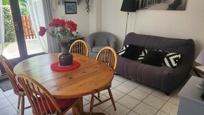 A dining area with a wooden table, chairs, a vase with red flowers, a grey sofa with patterned cushions, and a tiled floor.