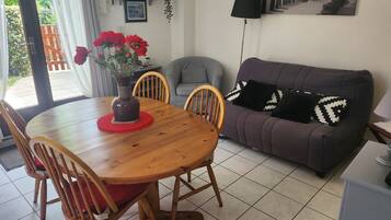 Een eetgedeelte met een houten tafel, stoelen, een vaas met rode bloemen, een grijze bank met gekleurde kussens, en een betegelde vloer.