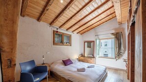 A bedroom with a wooden ceiling, a bed with white linens, a blue chair, and a window with a view of buildings. - Spacious loft on pedestrian street with views (Chamonix-Mont-Blanc)