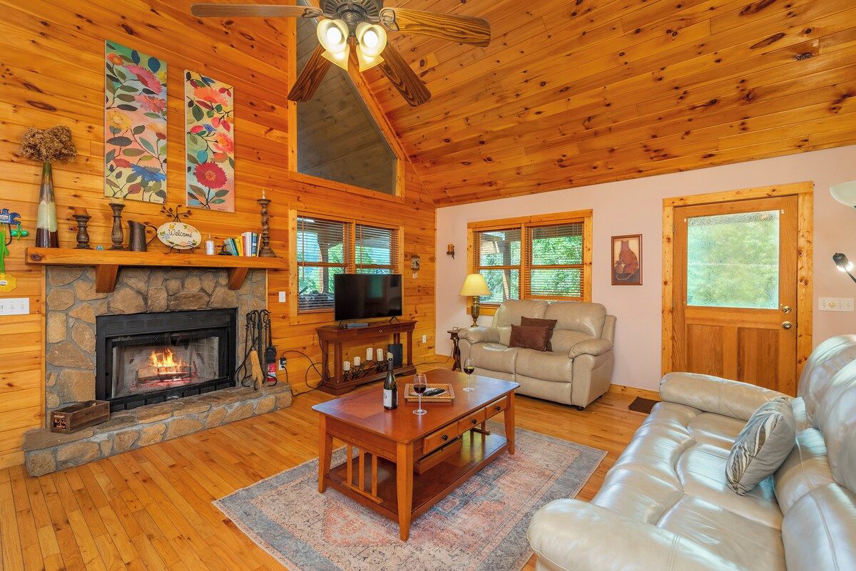 A cozy cabin interior with a stone fireplace, wooden walls, a leather sofa, and a television.