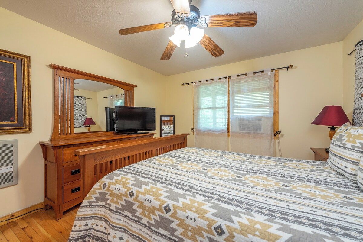 Une chambre à coucher comprenant un lit, une télévision, un ventilateur de plafond, une commode et une fenêtre avec des rideaux.