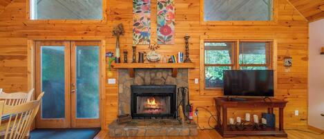 A log cabin interior with a stone fireplace, wooden walls, a television, and a dining area.