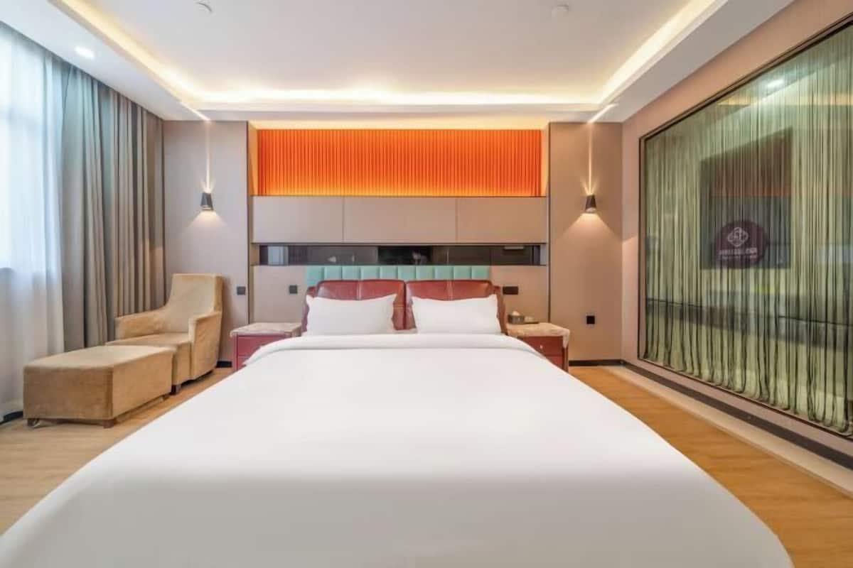 A modern hotel room with a large bed, a seating area with a chair and ottoman, a built-in TV, and a large window with curtains.
