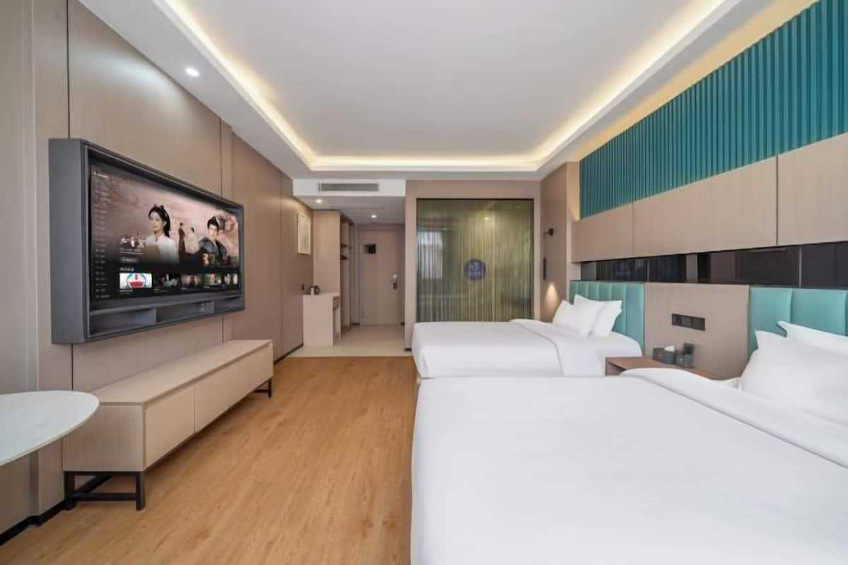 A modern hotel room with a large flat-screen TV, two beds, a seating area, and a bathroom visible through a glass door.
