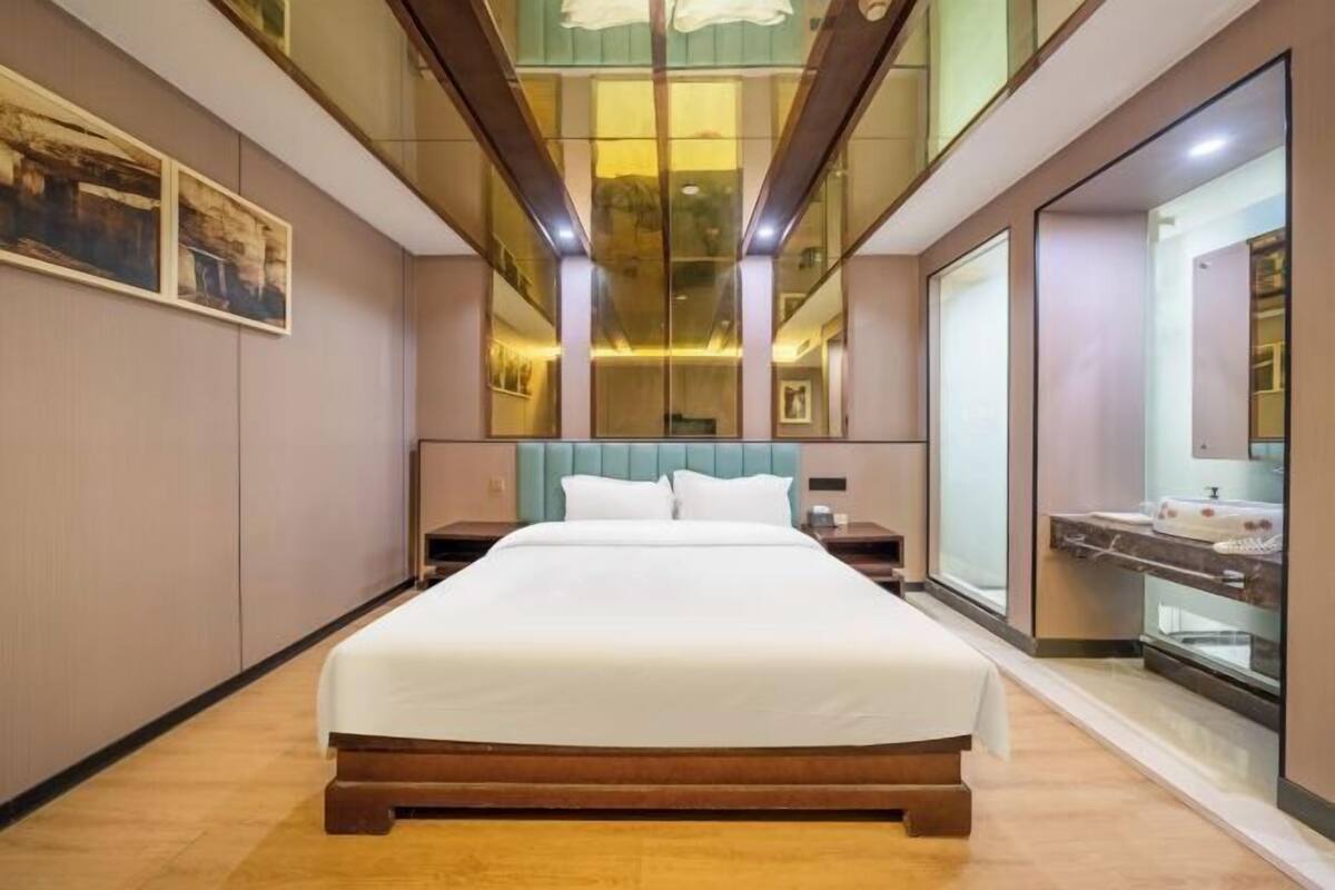 A modern hotel room with a large bed, a nightstand, a mirror, and a view of the city through large windows.