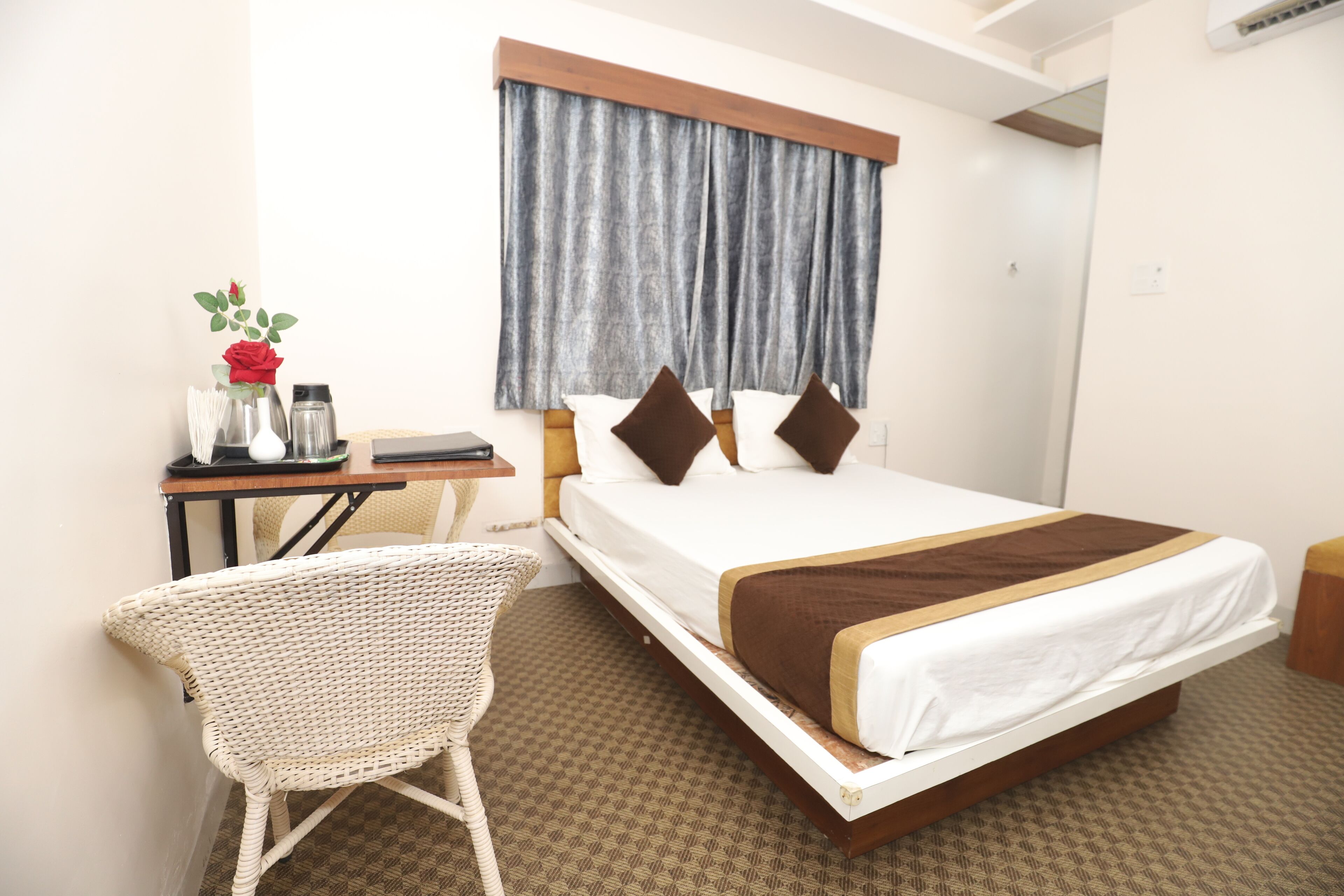 A hotel room with a bed, a desk with a chair, a vase with flowers, and a window with curtains.