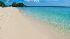 Une plage à l’eau turquoise et limpide, avec un rivage sablonneux et une végétation luxuriante en arrière-plan.