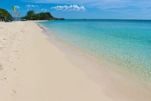 Une plage à l’eau turquoise et limpide, avec un rivage sablonneux et une végétation luxuriante en arrière-plan.