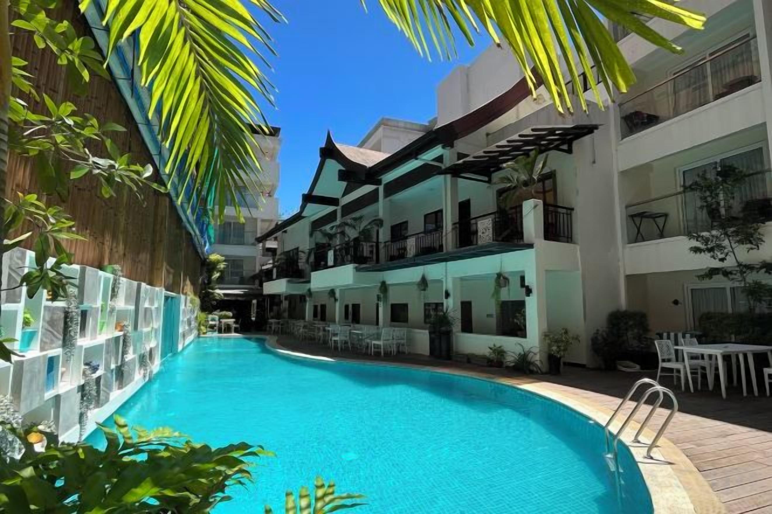 A swimming pool surrounded by multi-story buildings with balconies, outdoor seating, and tropical plants.