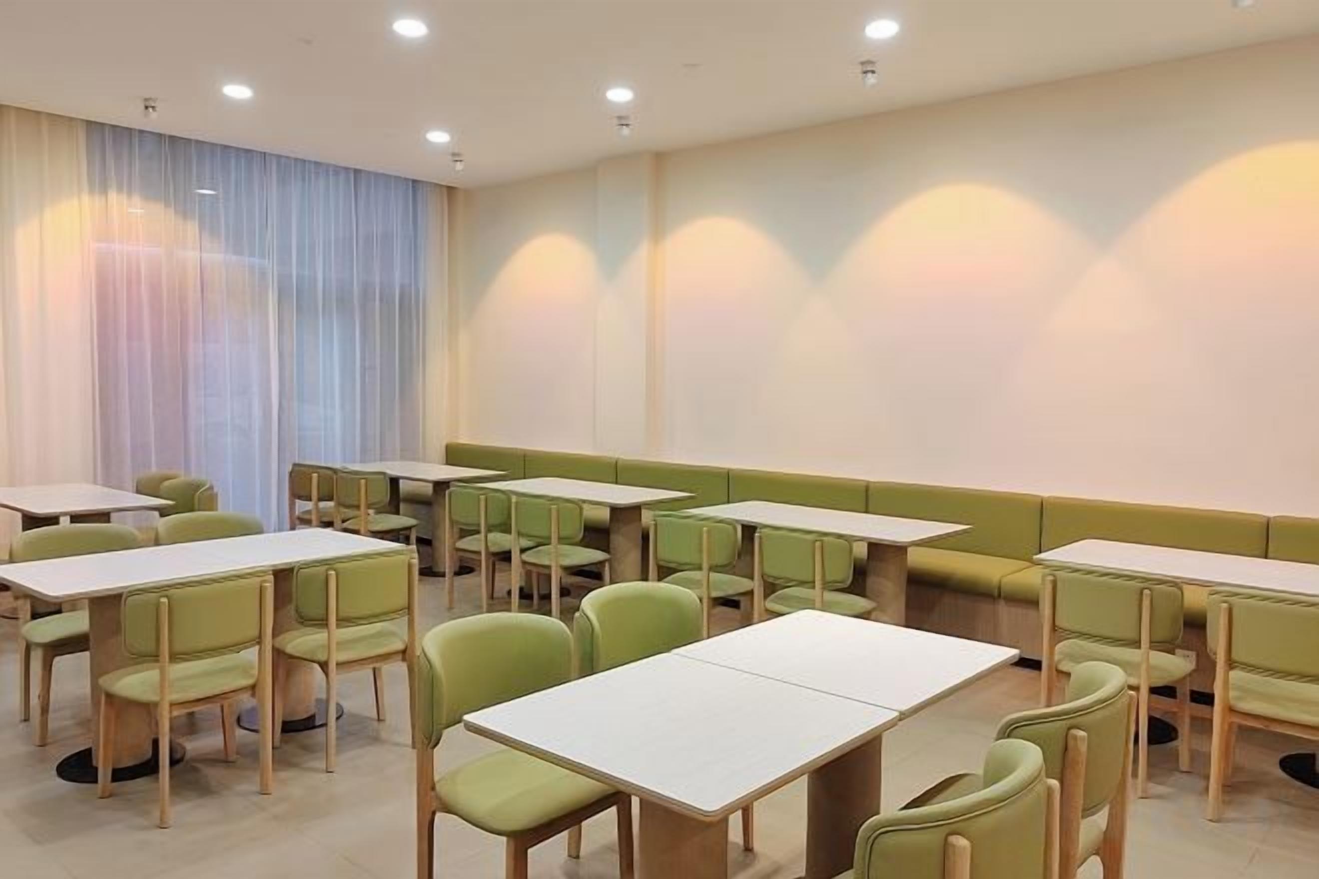 A dining area with tables and chairs arranged in rows, a large window with curtains, and recessed ceiling lighting.